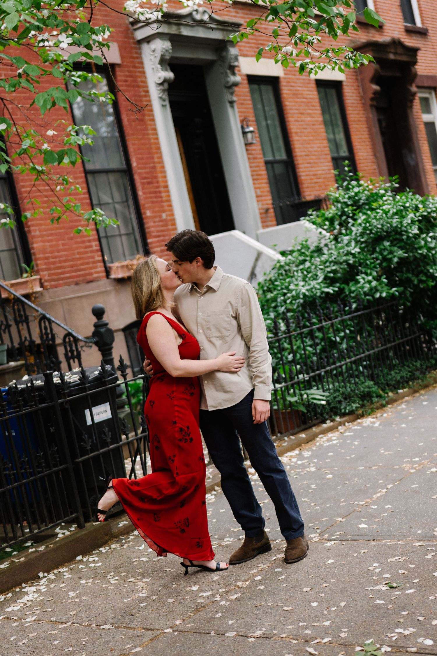 Couple sharing a romantic kiss on a city sidewalk, woman in red dress, brick townhouse backdrop.