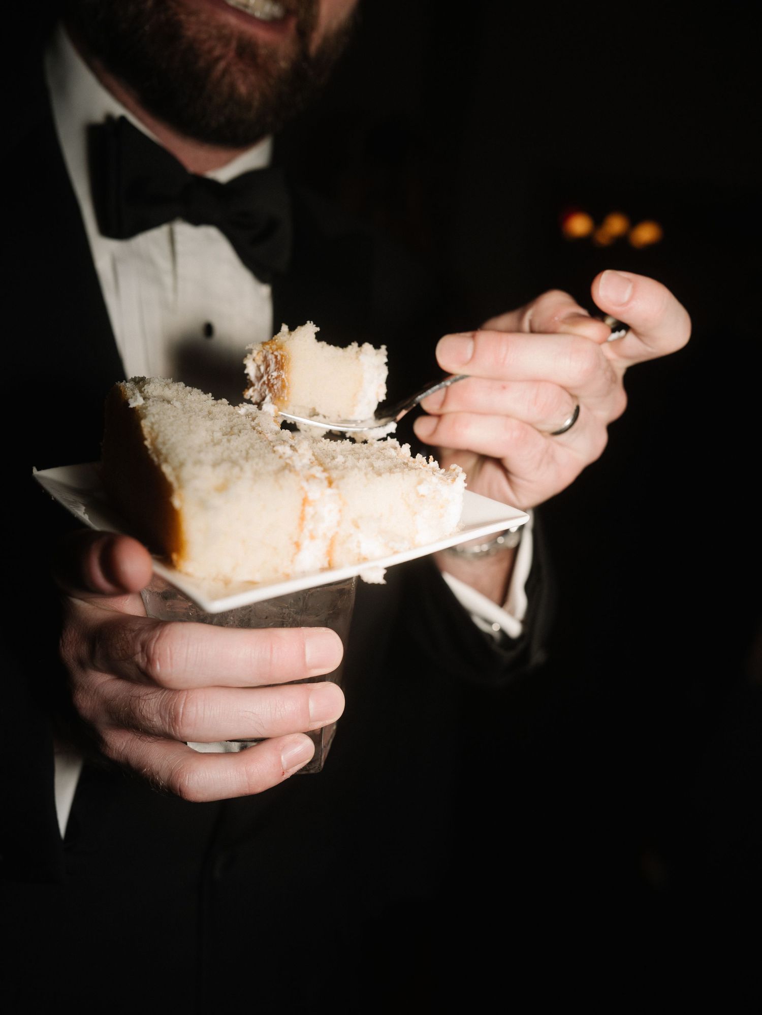 Groom in tuxedo with bow tie eating a slice of white wedding cake at a formal reception.