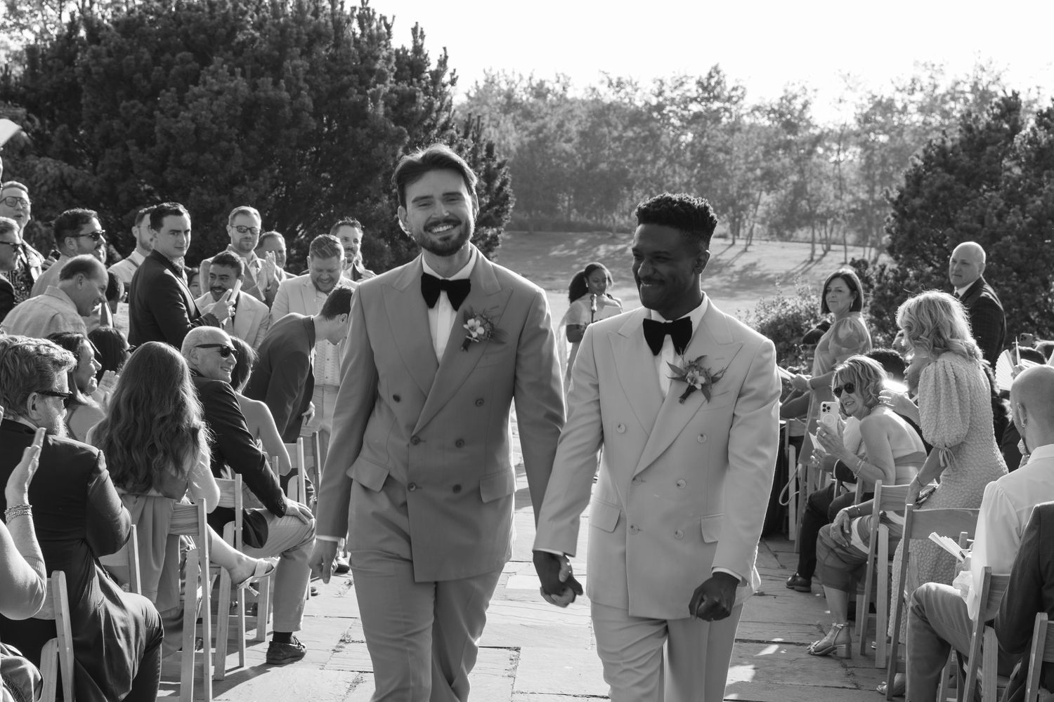 Two grooms walk hand-in-hand down the aisle at their outdoor wedding ceremony in black and white photo.