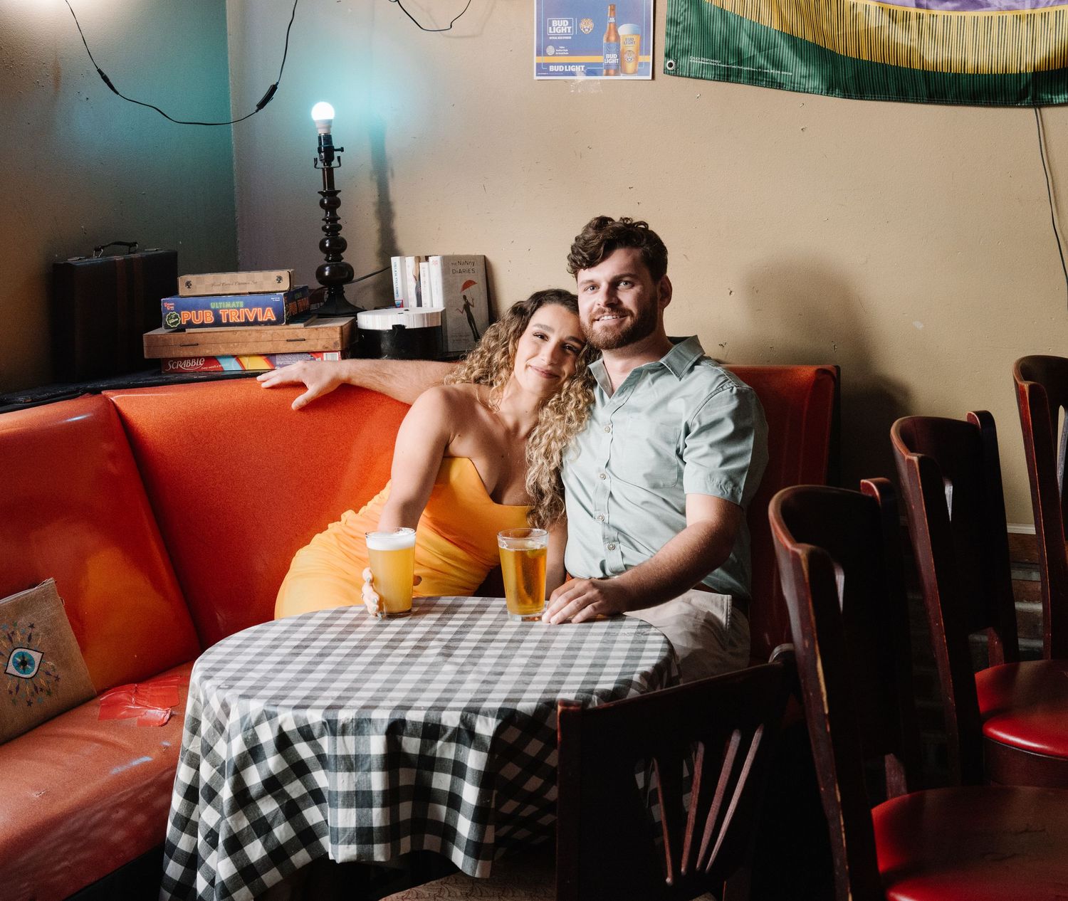 Couple sitting together on a red booth at a bar, smiling and holding drinks at a checkered table.