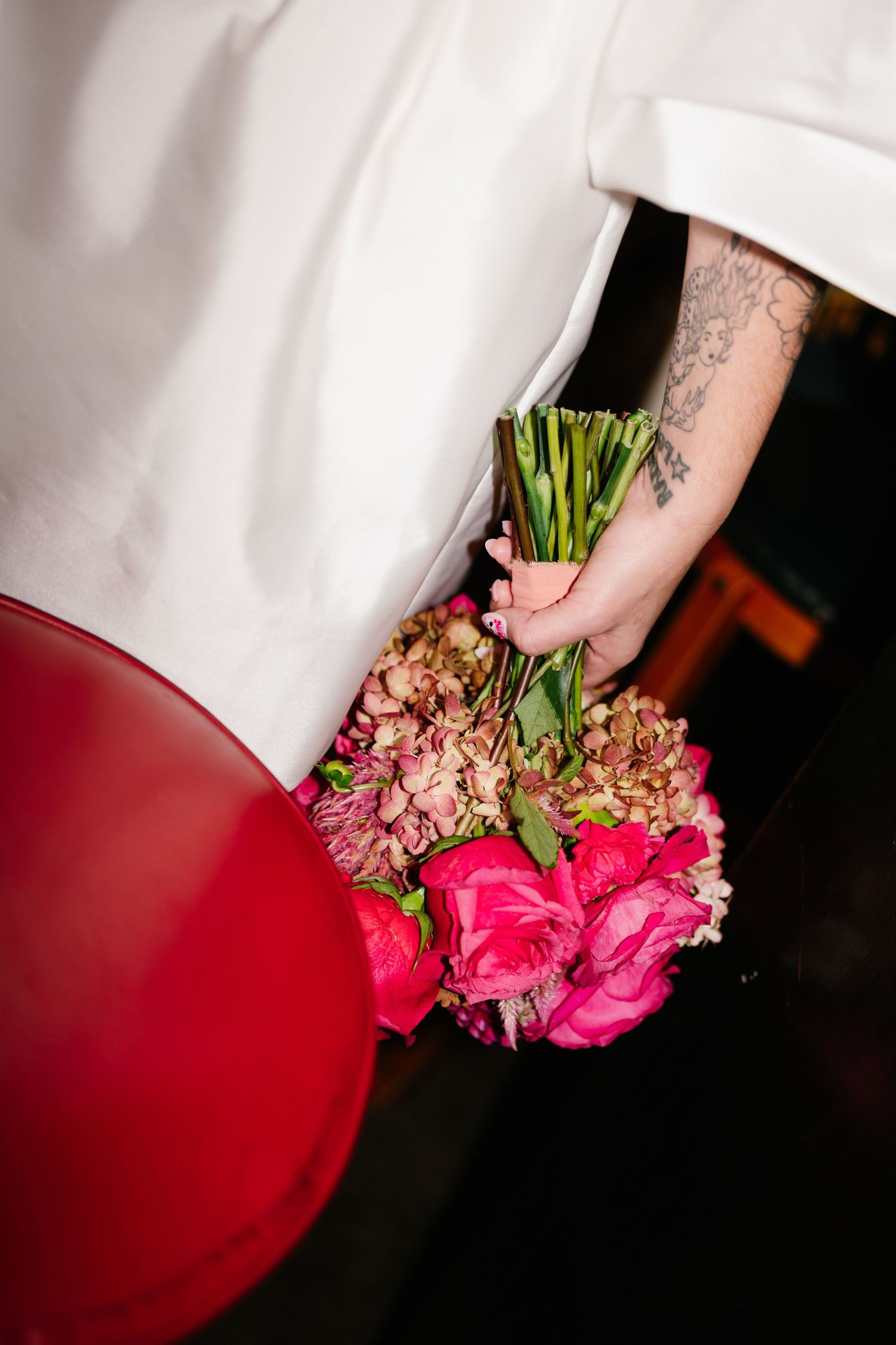 Bride's hand holding a vibrant bouquet of pink flowers and hydrangeas next to a red bar stool.