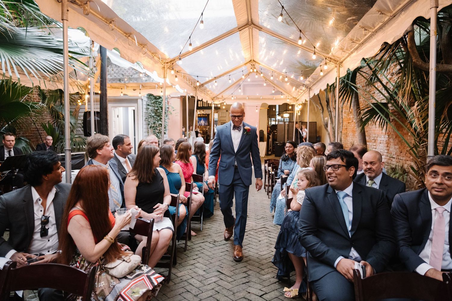 Elegant indoor wedding ceremony with guests seated along an aisle under string lights in a glass-roofed venue.