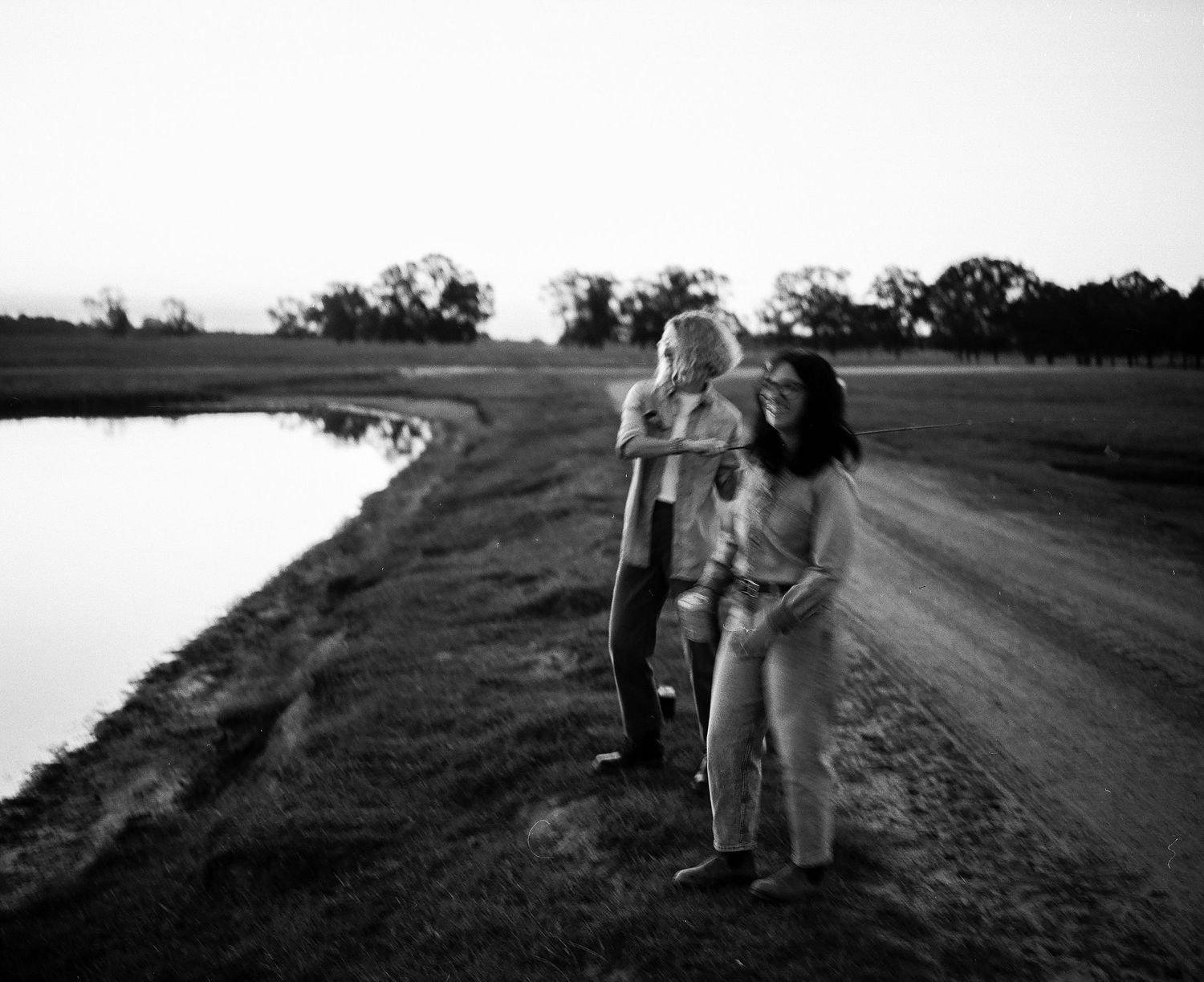 Black and white photo of a woman and child standing by a rural dirt road near a pond at sunset.