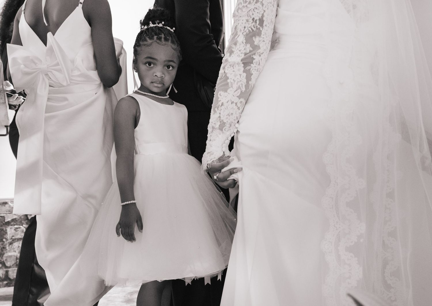 Black and white photo of a young flower girl in white dress standing among wedding gowns at a ceremony.