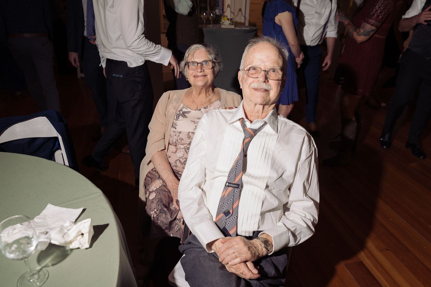 Elderly couple at a formal event, man in white shirt and tie seated, woman in beige top smiling behind him.