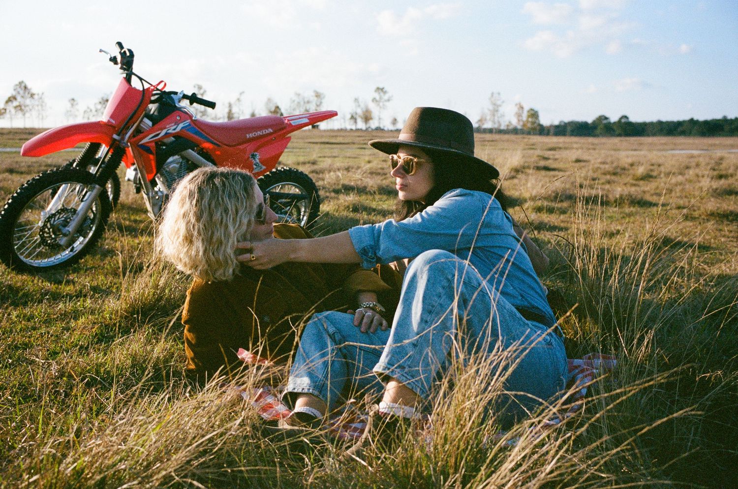 Two people sitting in a grassy field with a red dirt bike parked behind them on a sunny day.