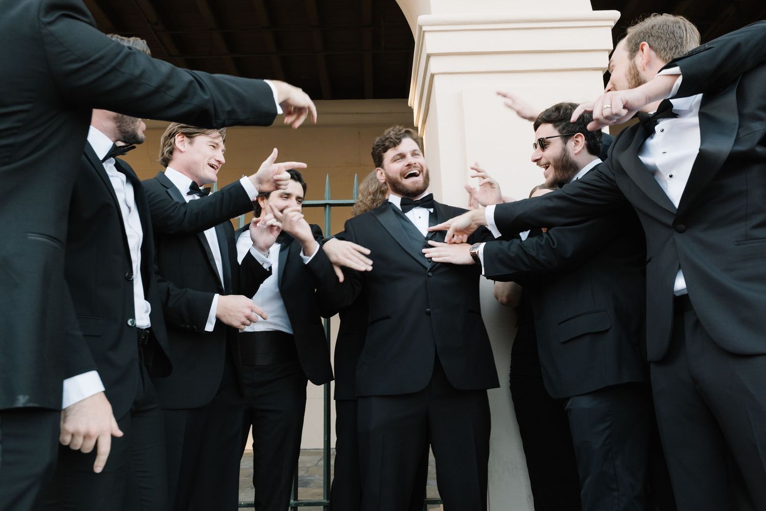 Groomsmen in black tuxedos laughing and celebrating together outdoors at a wedding reception.
