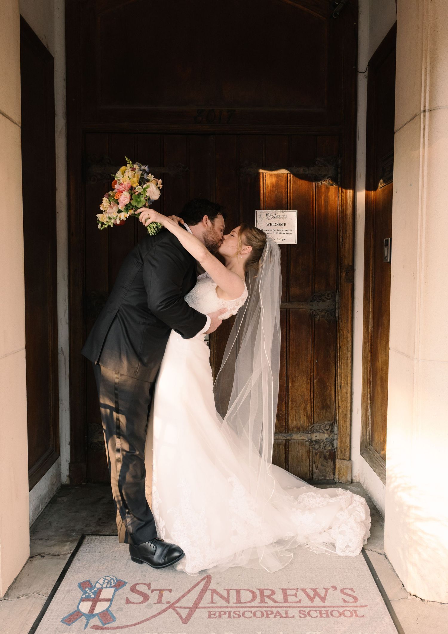 Newlyweds share a romantic kiss at St. Andrew's Episcopal School entrance, bride in white gown with veil holding colorful bouquet.