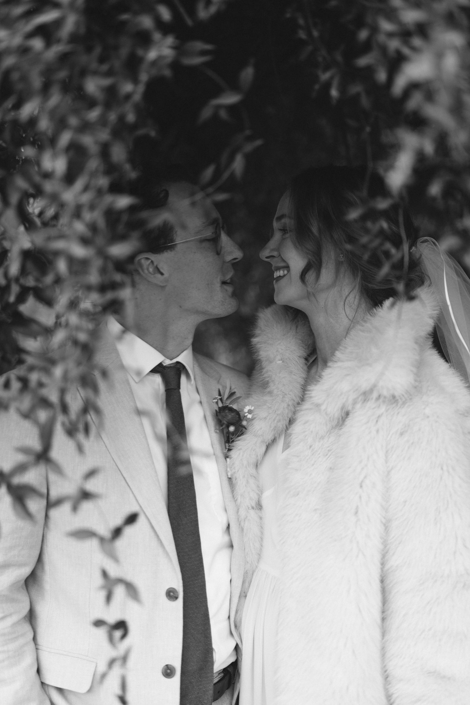 Black and white wedding photo of smiling bride in fur coat and groom in suit sharing a romantic moment among lush foliage.