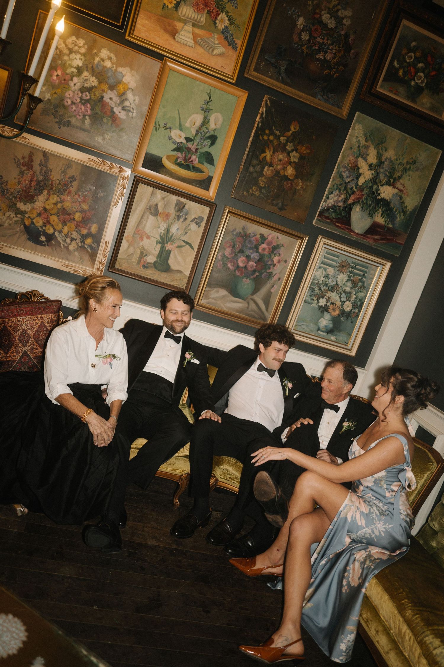Four elegantly dressed wedding guests sit and chat on a gold sofa surrounded by vintage floral oil paintings.