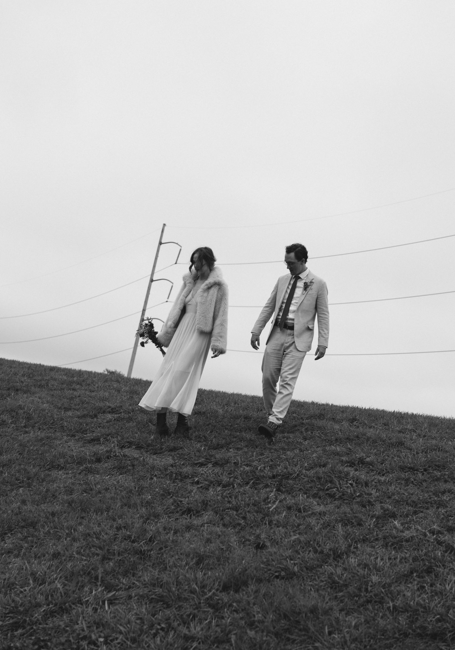 Black and white photo of a bride and groom walking hand in hand across a grassy hill on their wedding day.