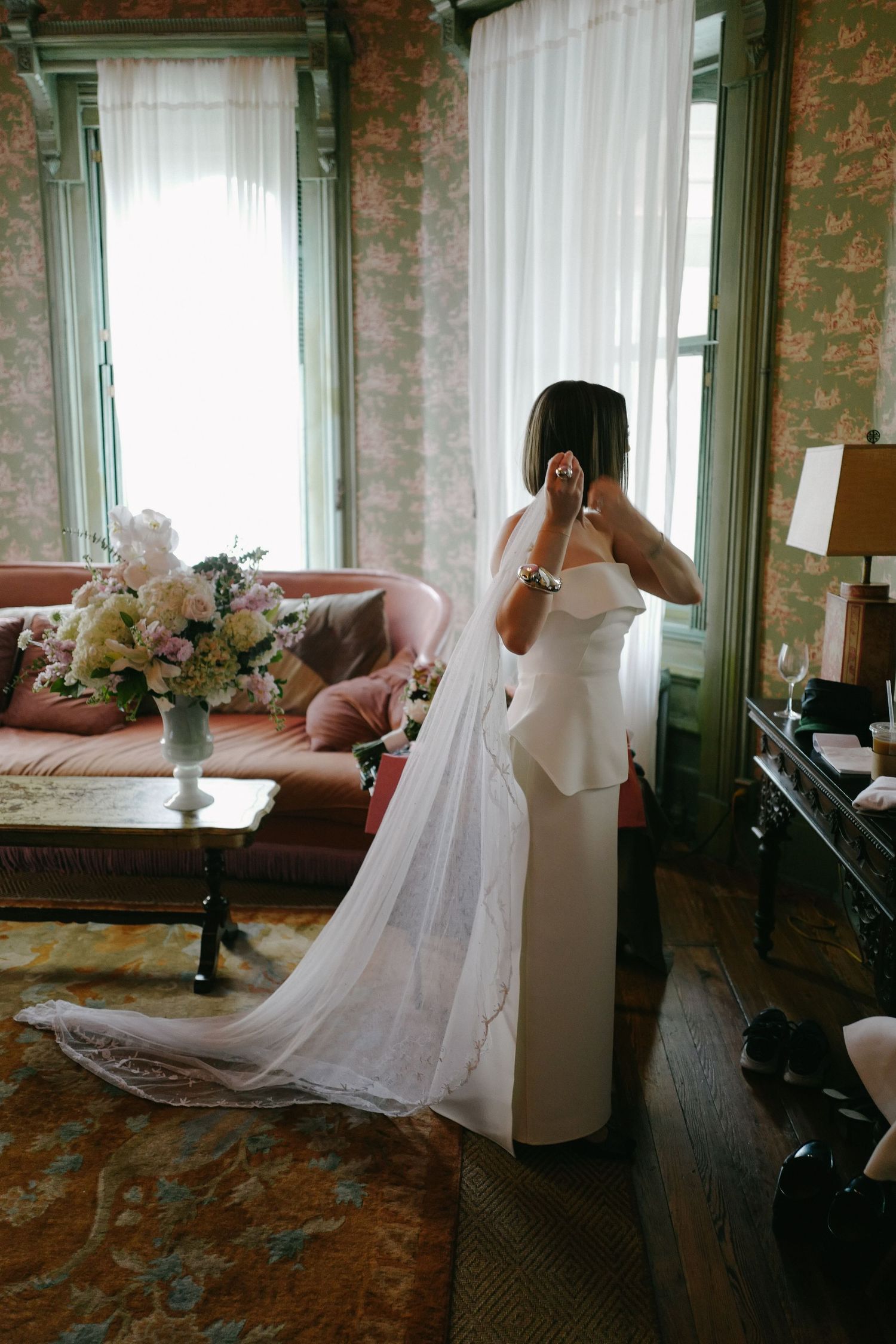 A bride in a flowing white wedding dress poses in a vintage room with floral wallpaper.