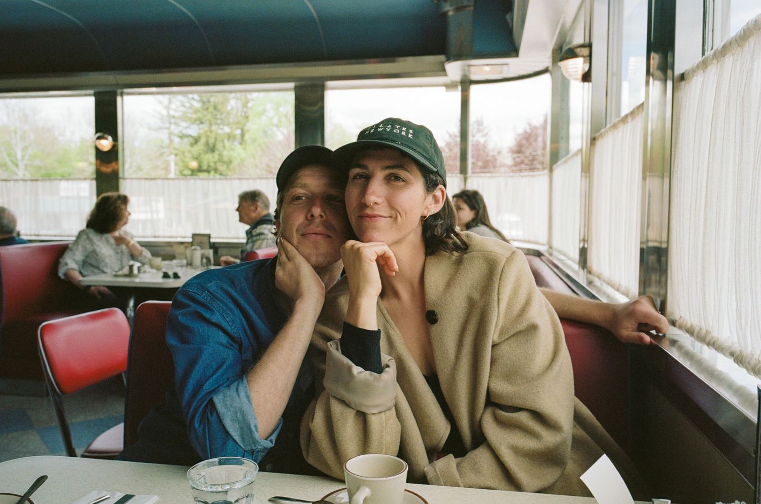 Two friends pose together at a retro diner booth, one wearing a green cap and beige coat.
