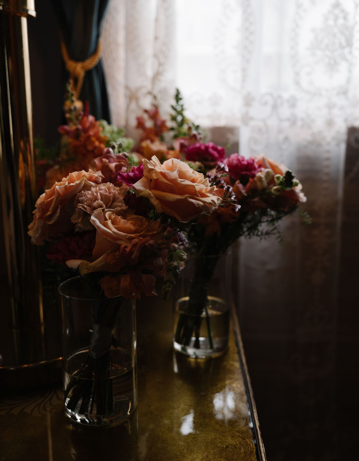 Two elegant floral bouquets with peach roses and pink blooms in glass vases on a reflective surface near a lace curtain.