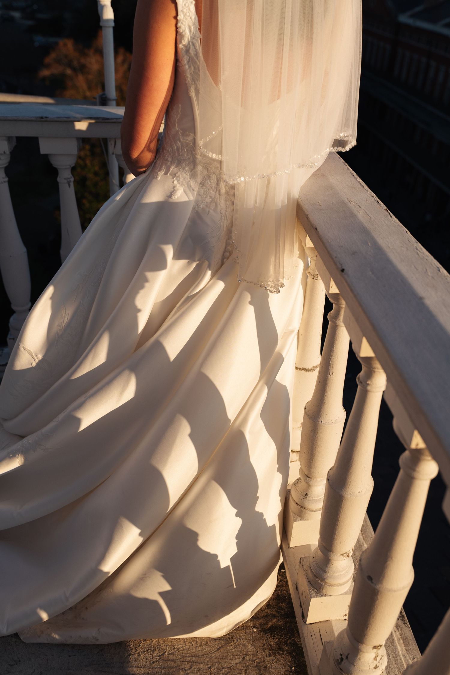 Bride in elegant white wedding gown with flowing skirt and veil standing on sunlit white balcony railing.