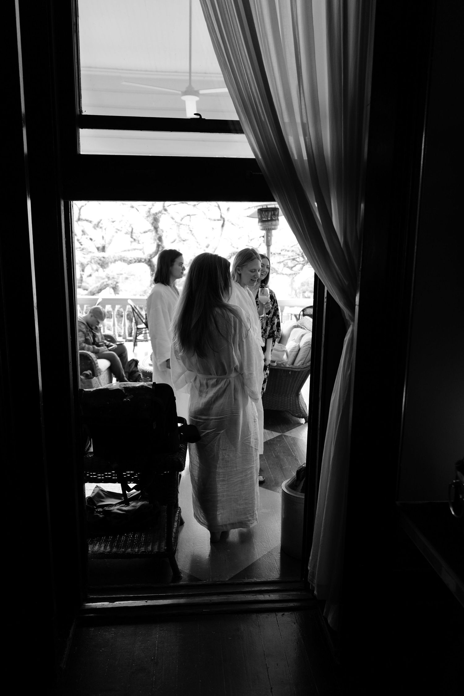 View through a doorway showing bridesmaids in white robes silhouetted against bright window light.