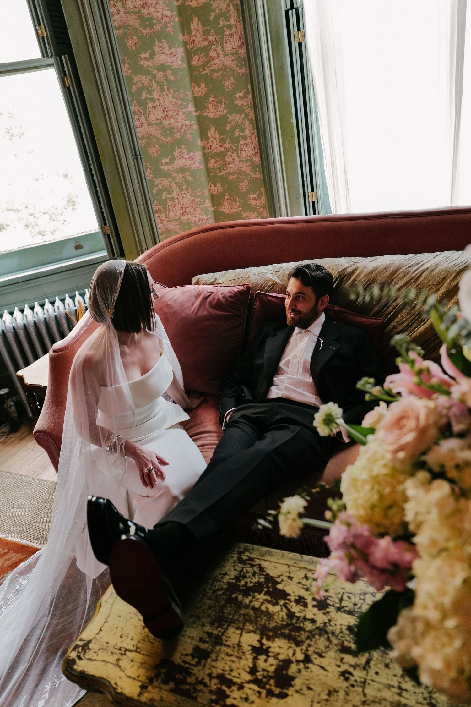 Romantic wedding couple shares an intimate moment on a red couch at The Columns Hotel in New Orleans surrounded by floral arrangements.