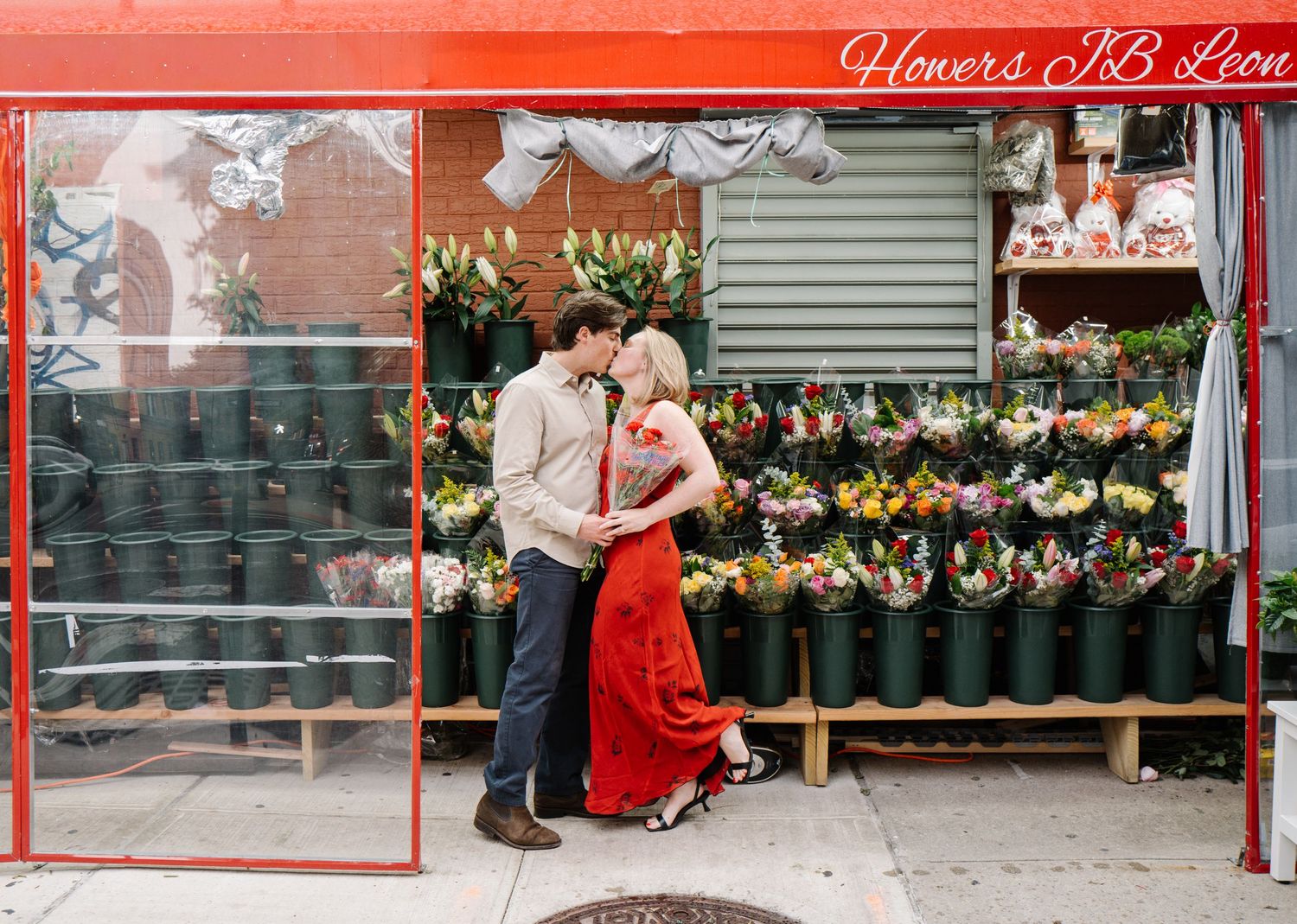 Couple kissing in front of a colorful flower shop with red framed windows and vibrant bouquets on display.