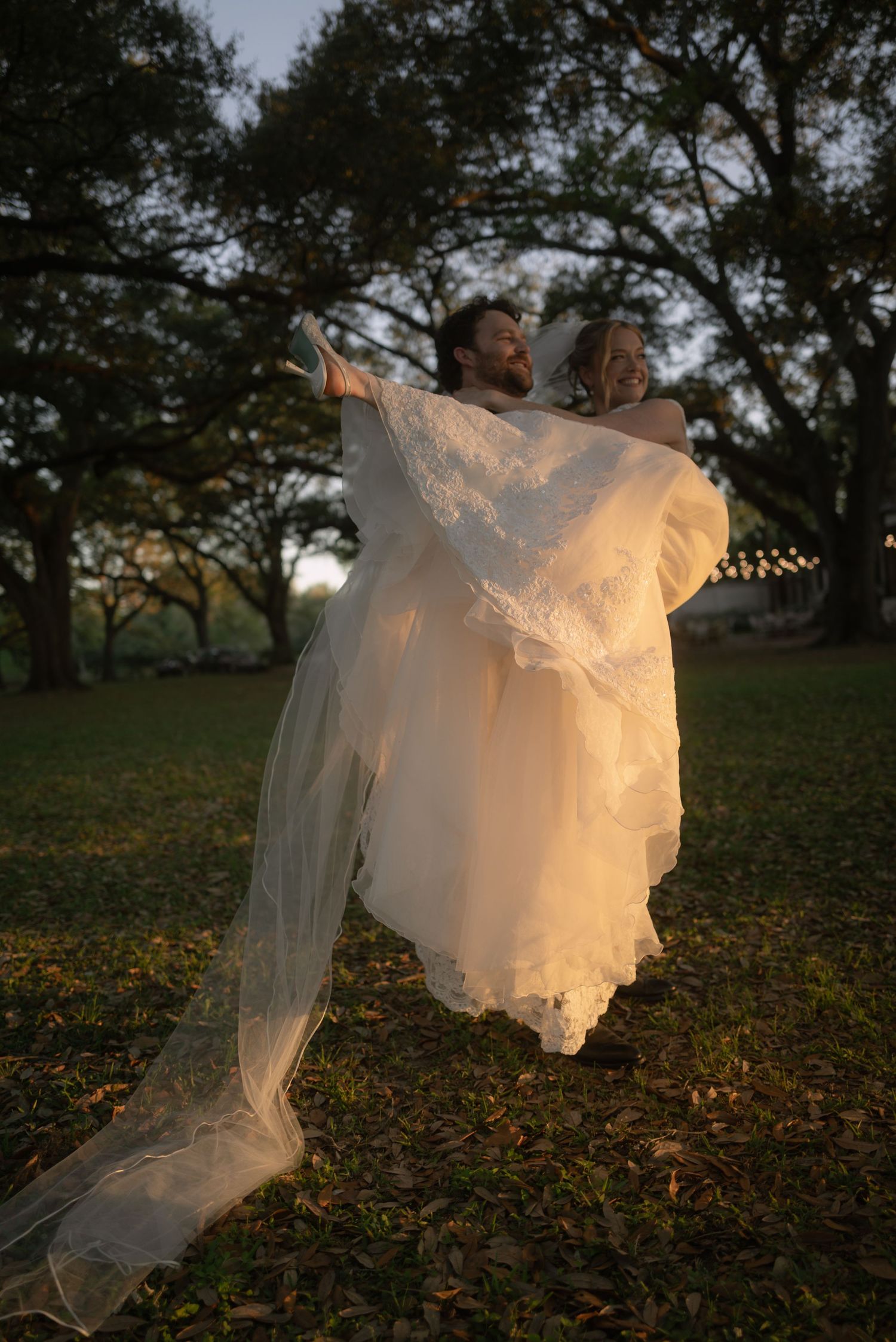 Groom lifting bride in white gown outdoors under oak trees at golden hour during wedding celebration.