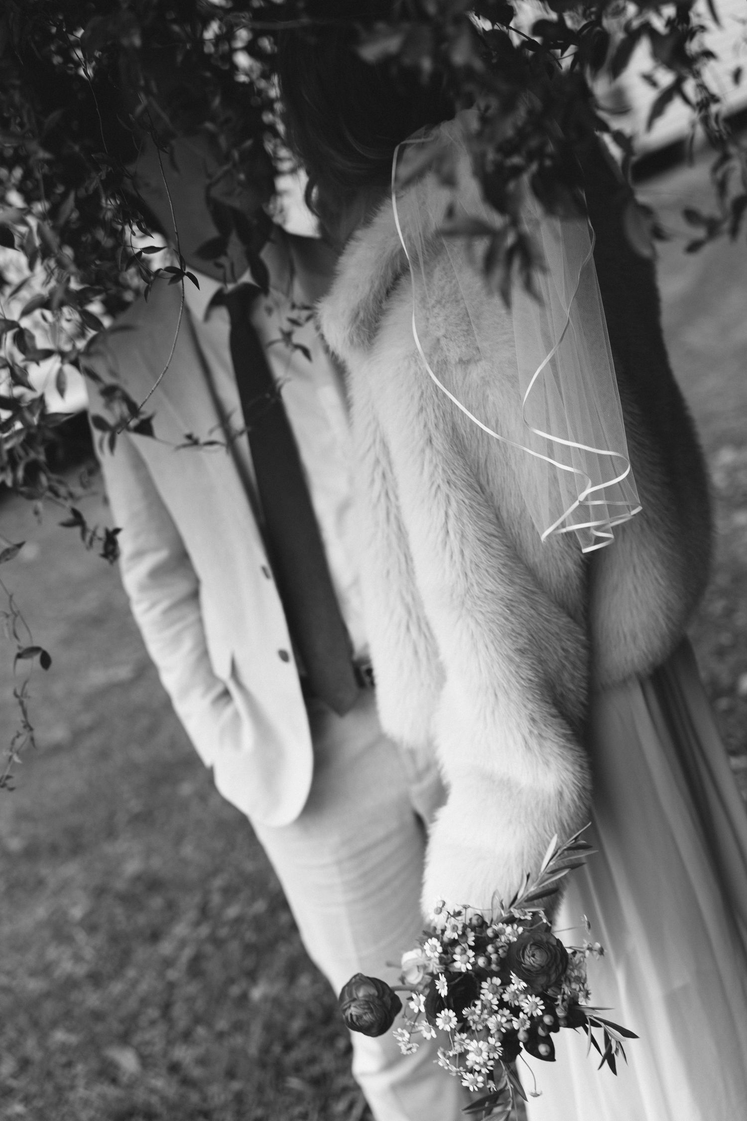 Black and white photo of bride in fur stole holding bouquet beside groom in suit under leafy branches.