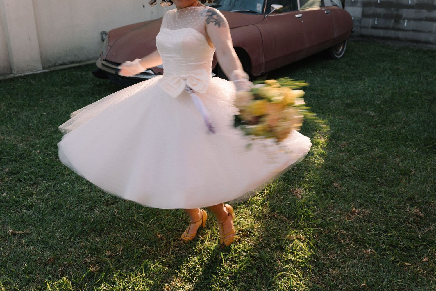 Bride in a white tea-length tulle wedding dress twirling outdoors, holding yellow bouquet, with a vintage pink car in background.