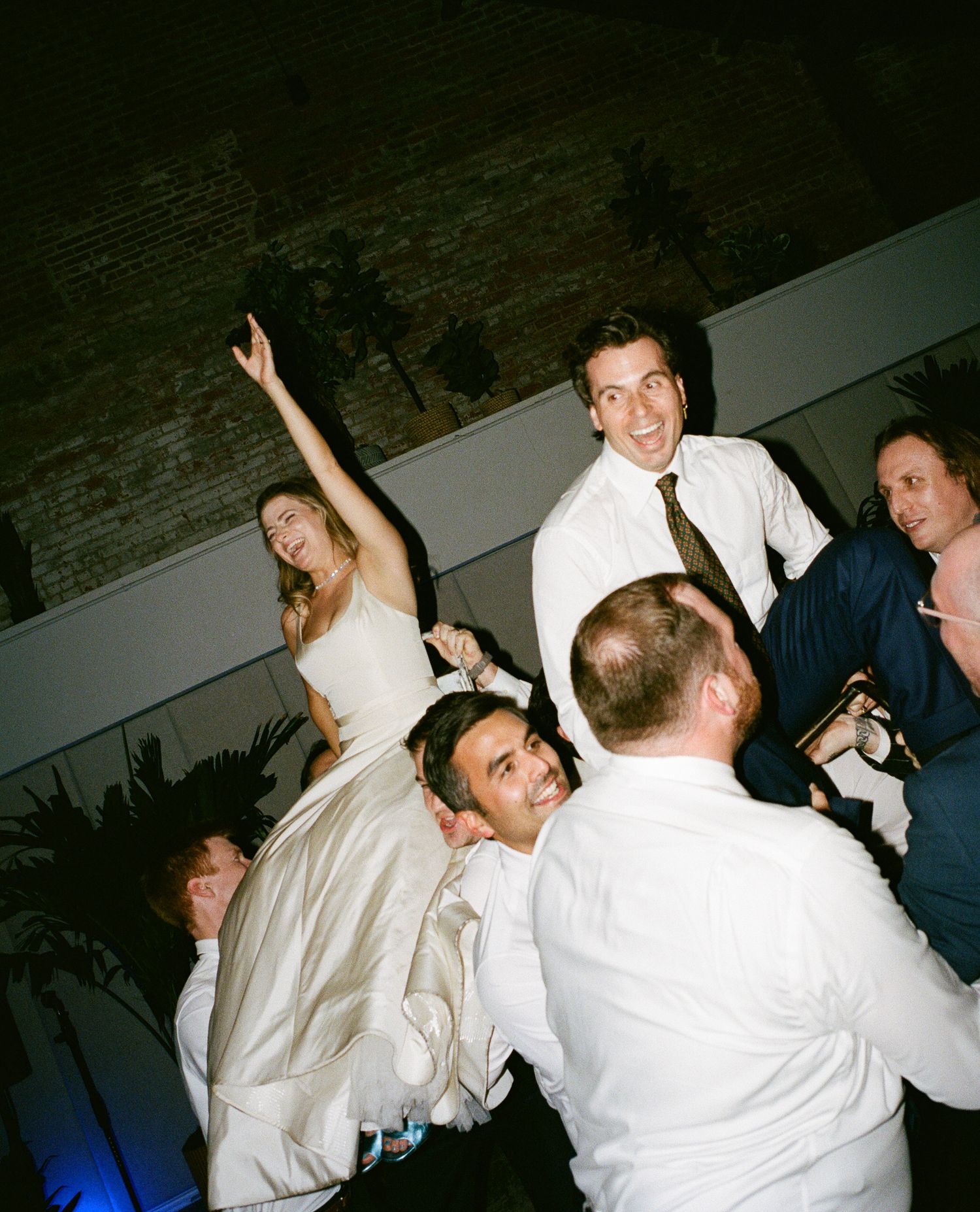 A joyful wedding reception scene with bride in white dress raised up by guests while groom celebrates nearby indoors.