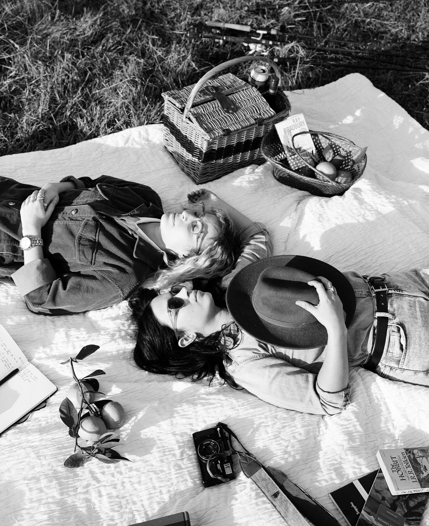 Vintage black and white photo of a couple lying on a picnic blanket outdoors with baskets and a camera nearby.
