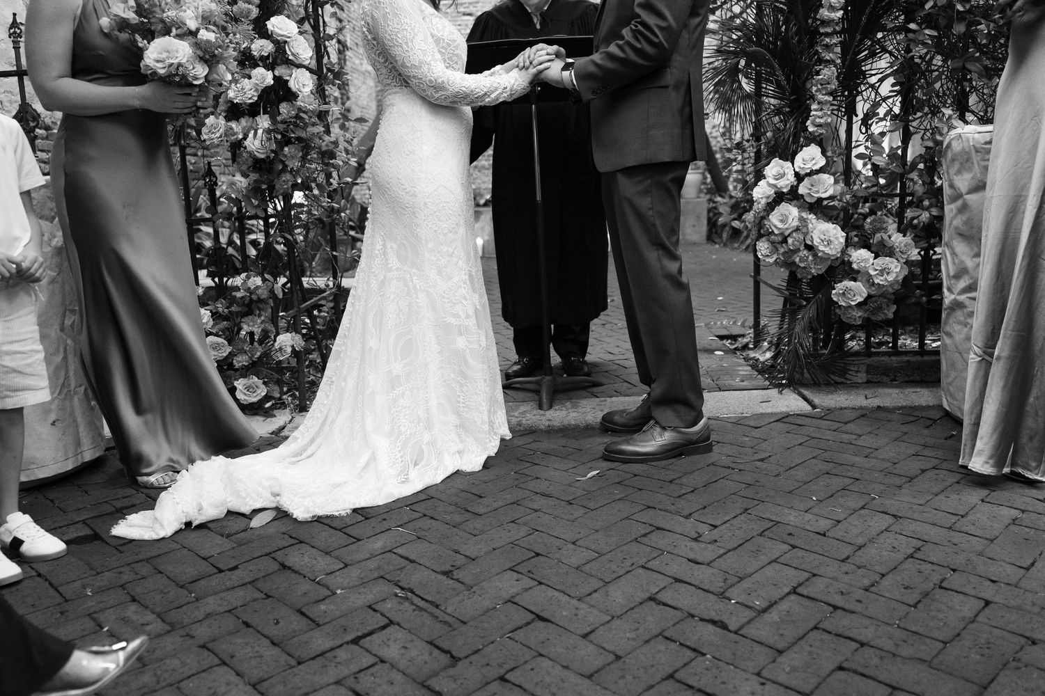 Black and white wedding ceremony photo showing bride in lace gown and groom exchanging vows on brick patio.