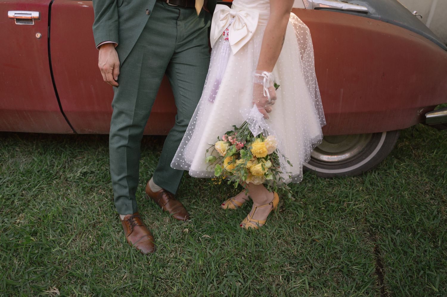 Bride and groom standing by vintage car, showing green suit, brown shoes, white tulle dress, and yellow floral bouquet.