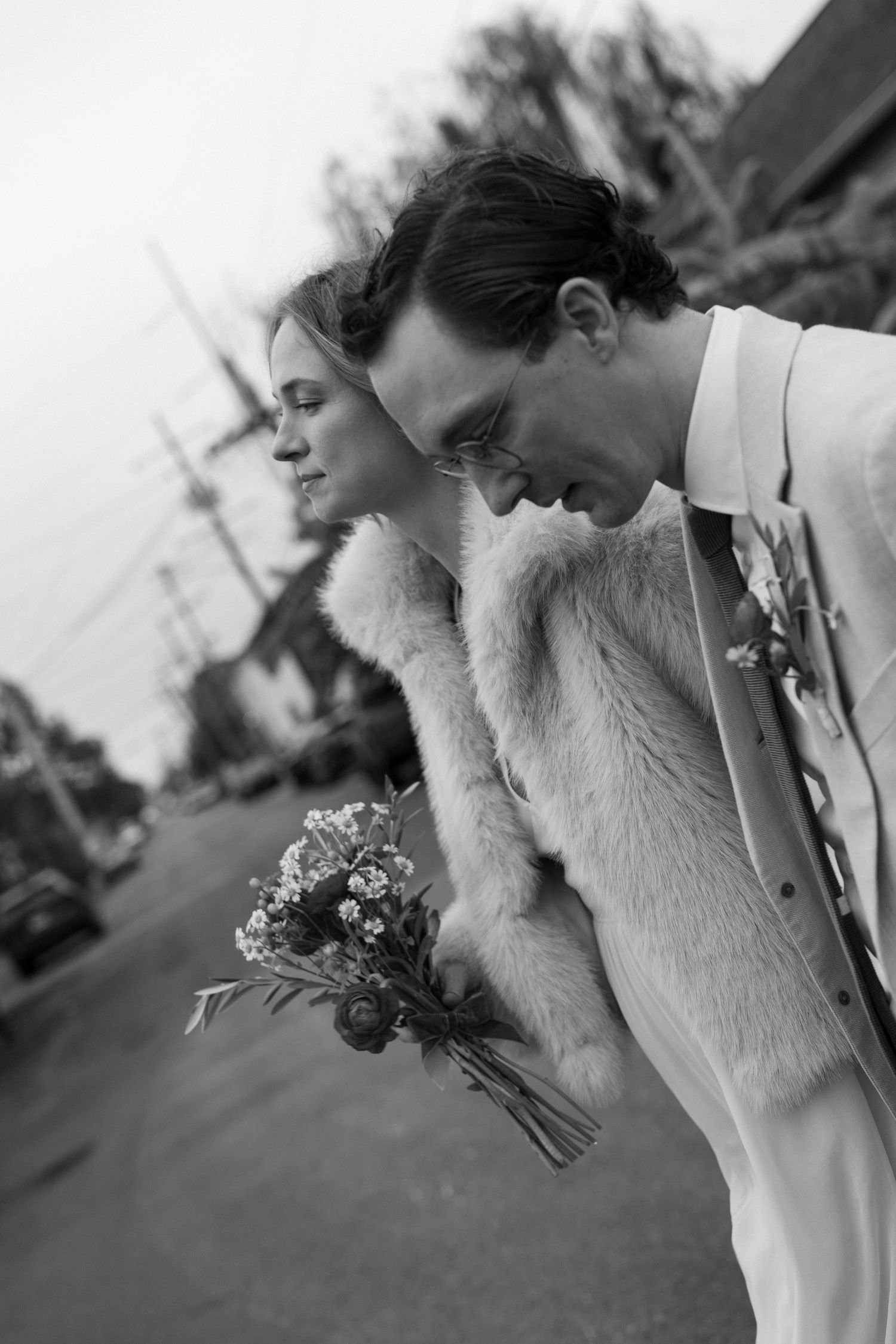 Black and white photo of a bride and groom leaning together, she holds a wildflower bouquet, wearing a fur stole.