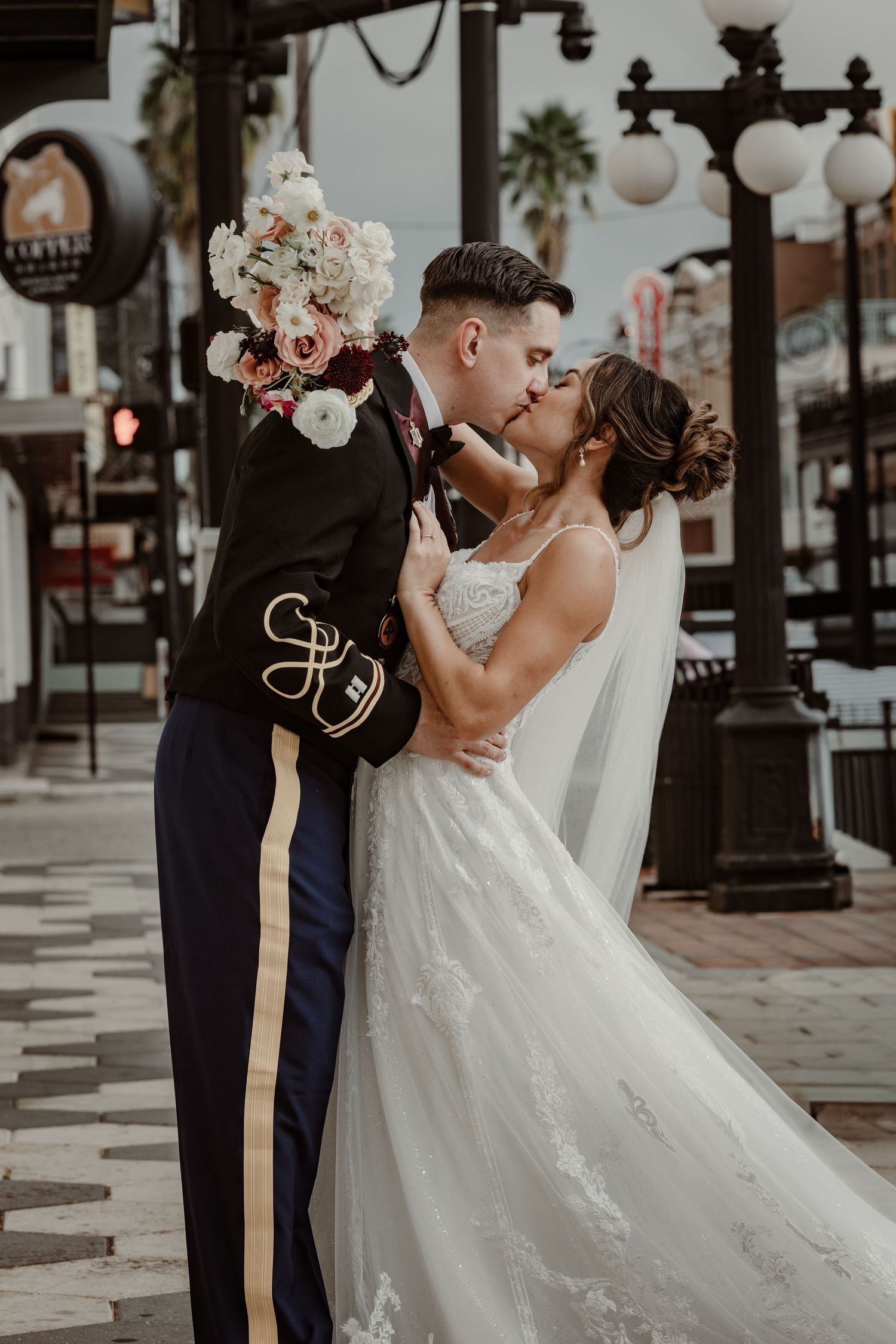 A wedding couple shares a romantic moment on a city sidewalk with decorative lamp posts.