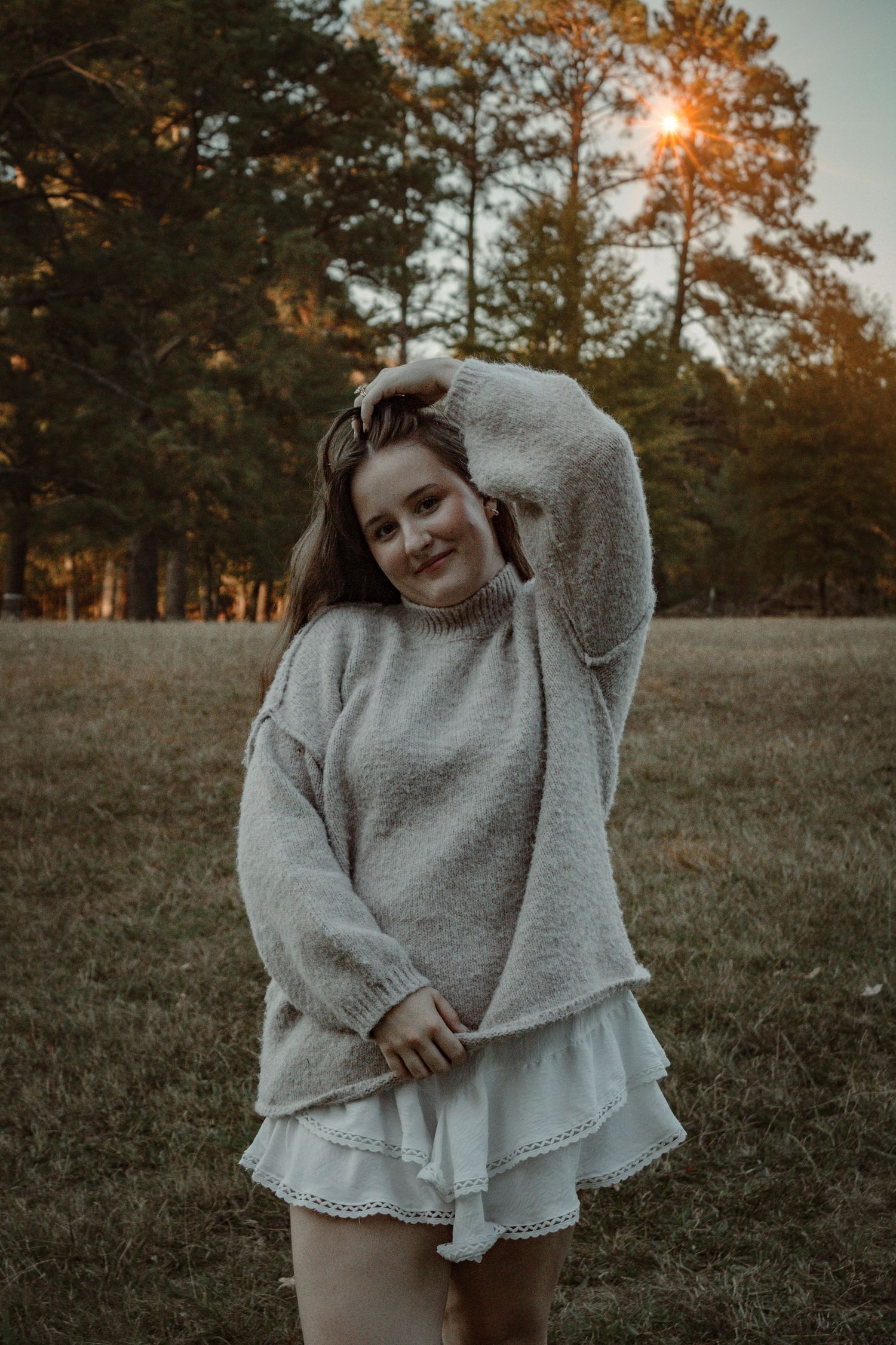 A person wearing a cozy gray sweater and white skirt twirls in a sunlit autumn field at golden hour.