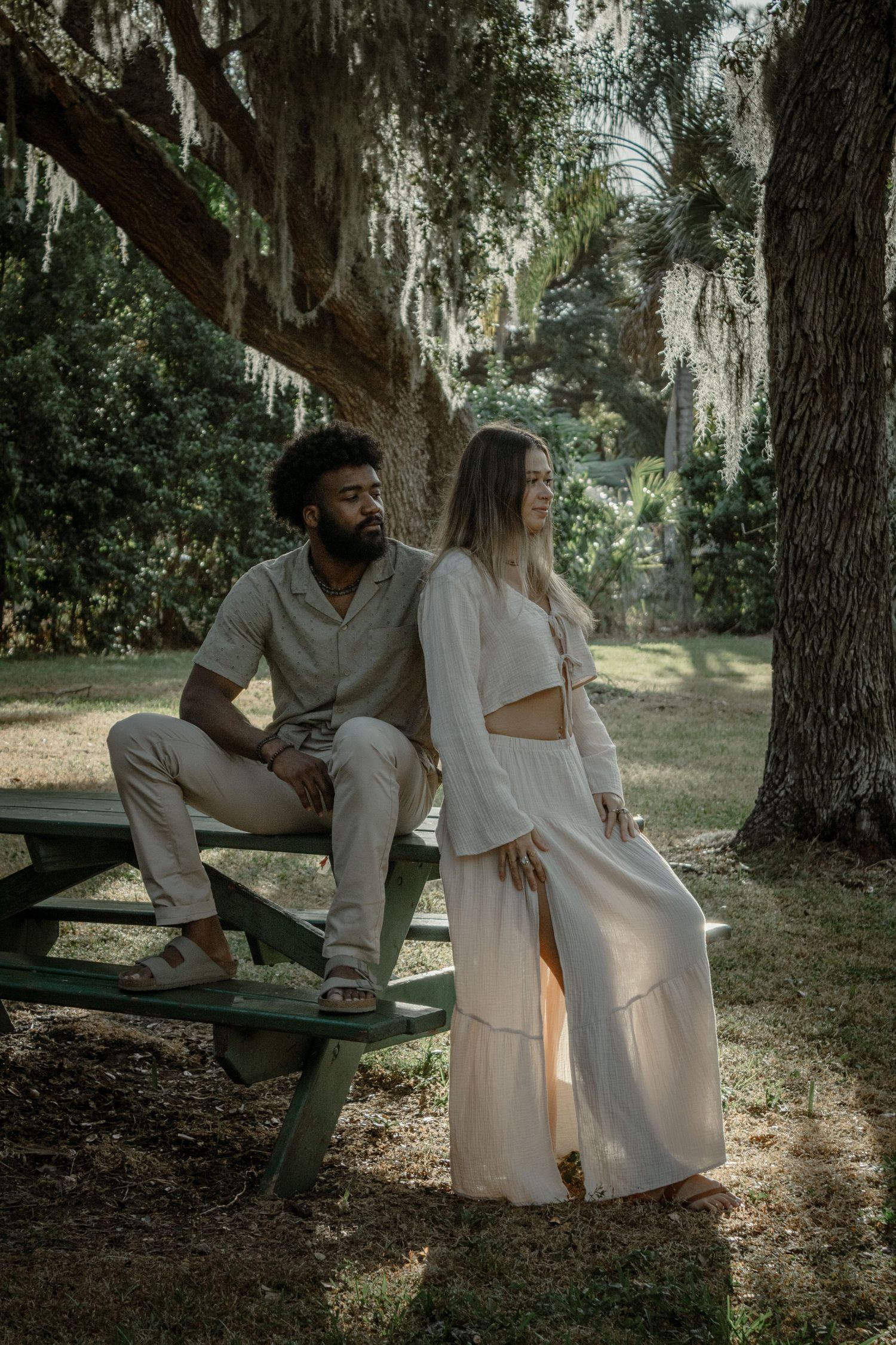 A romantic couple poses by a picnic table under moss-draped trees in a dreamy outdoor setting.