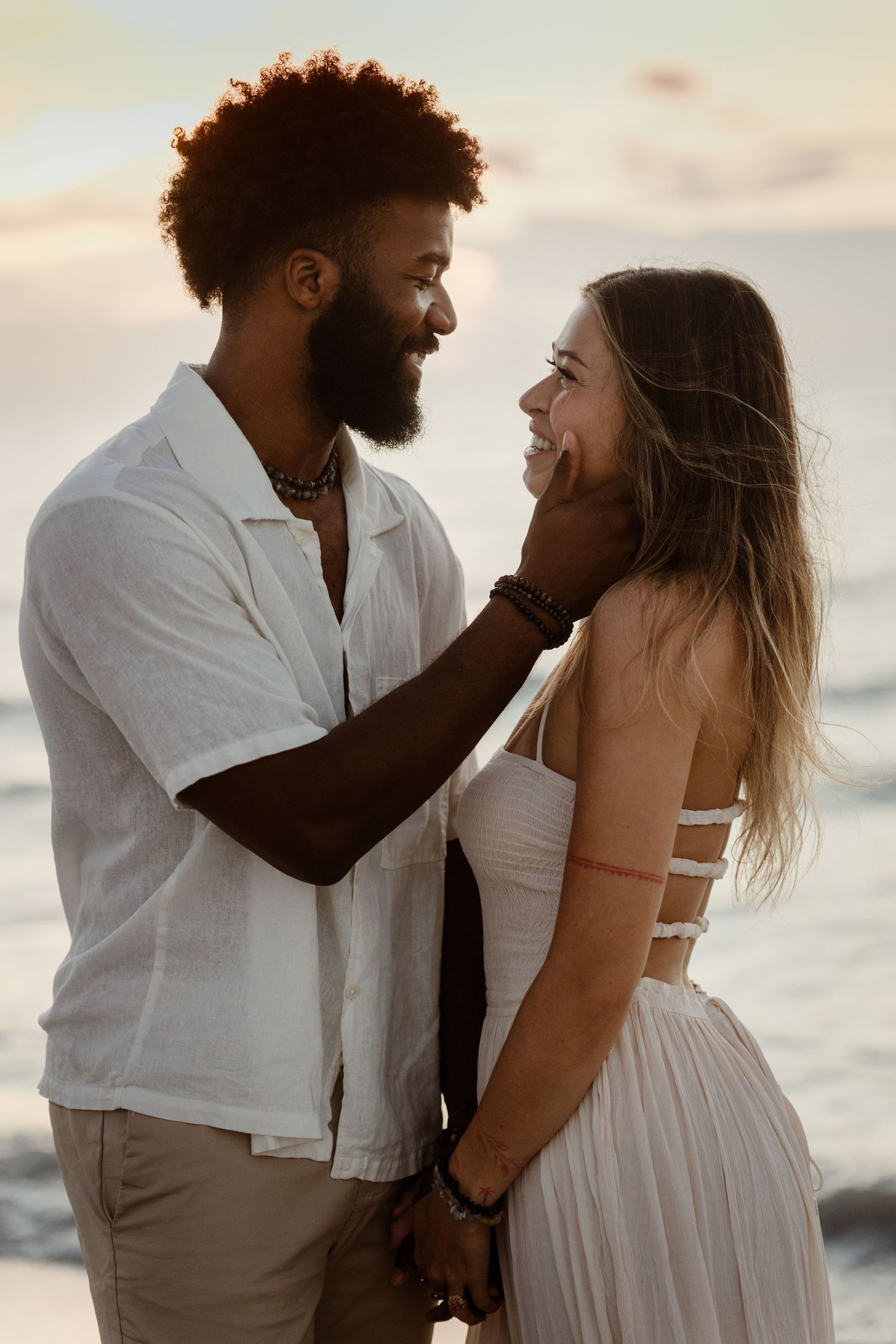 Couple in white outfits embraces during sunset photoshoot at the beach.