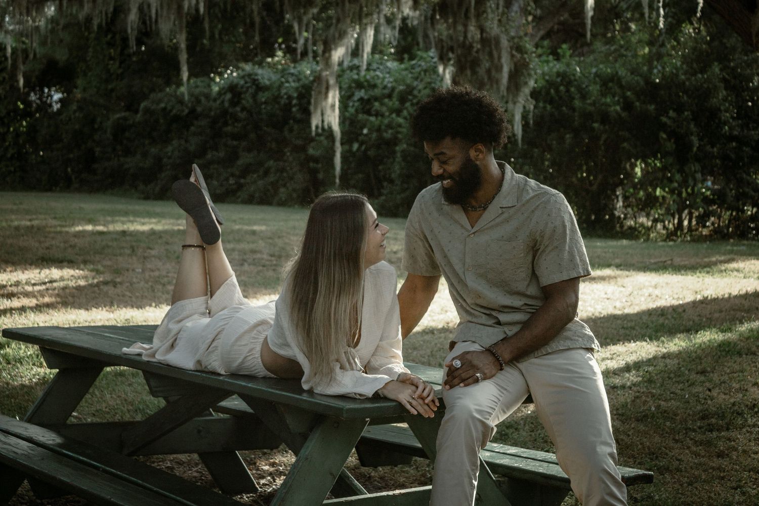 A couple sits together on blankets under moss-draped trees in a romantic outdoor setting.