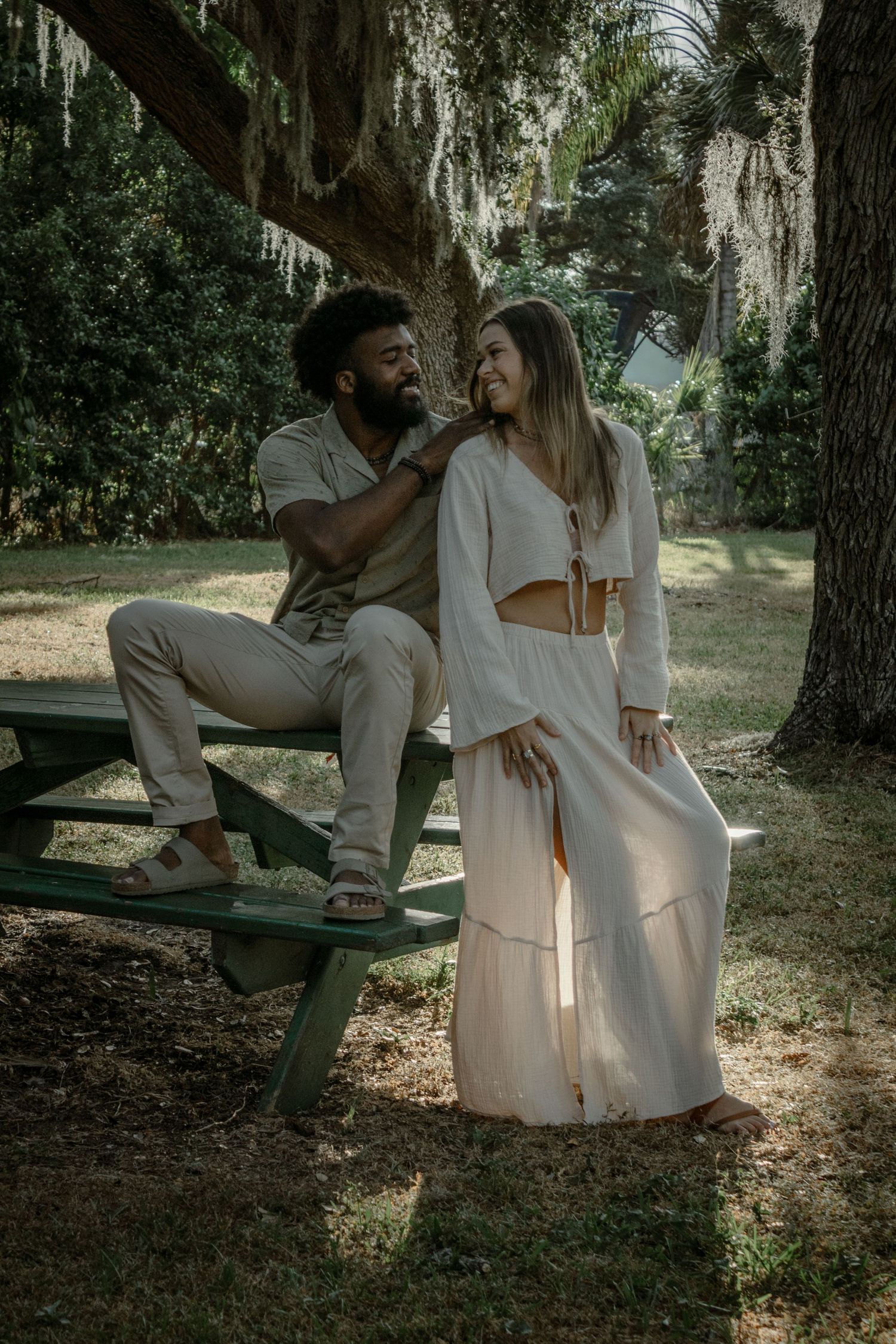 A romantic couple in white outfits sits together on a wooden bench under moss-draped trees in a dreamy garden setting.
