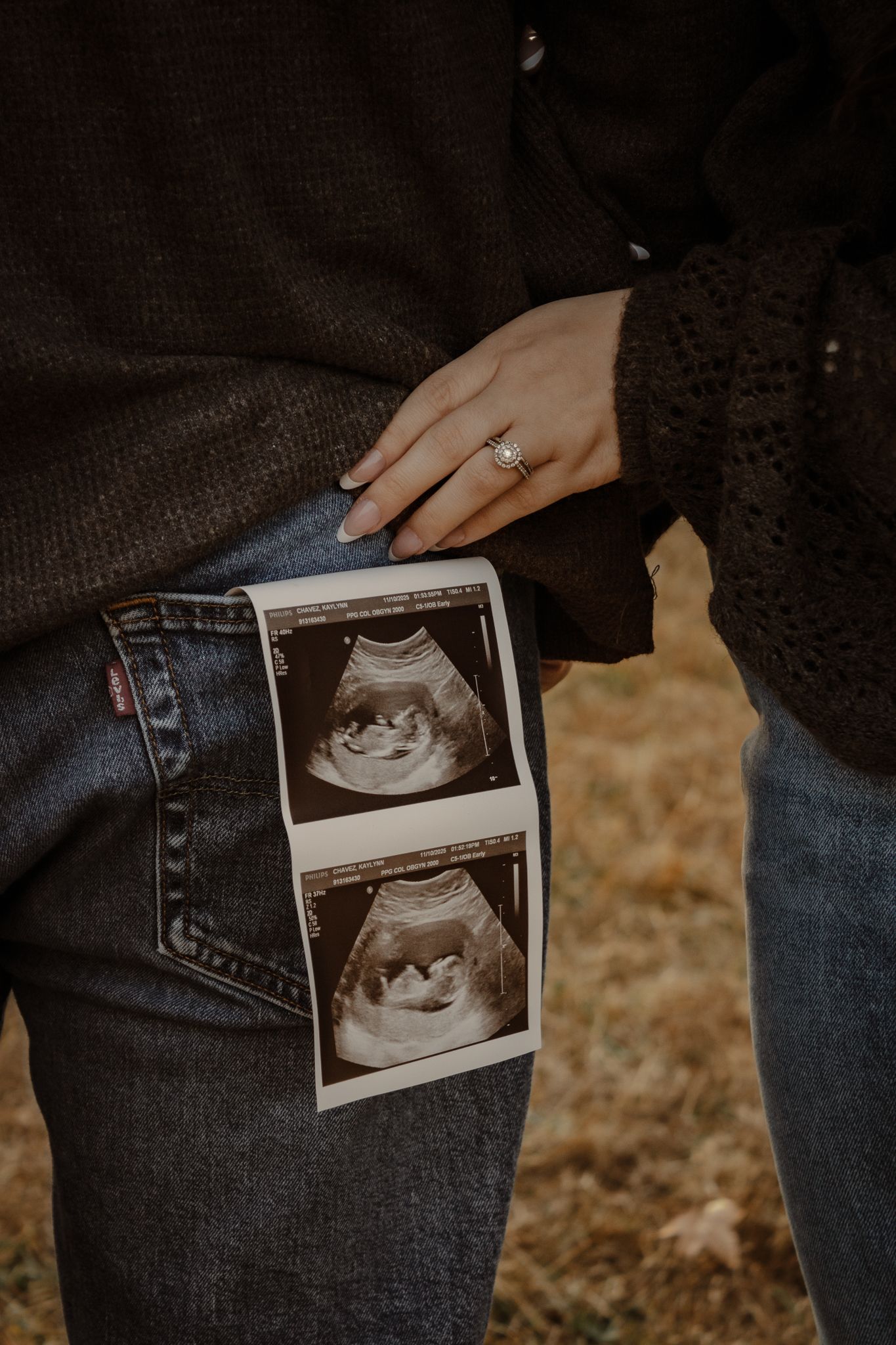 Hand wearing engagement ring holds ultrasound photos showing baby scans against denim jeans in outdoor setting.