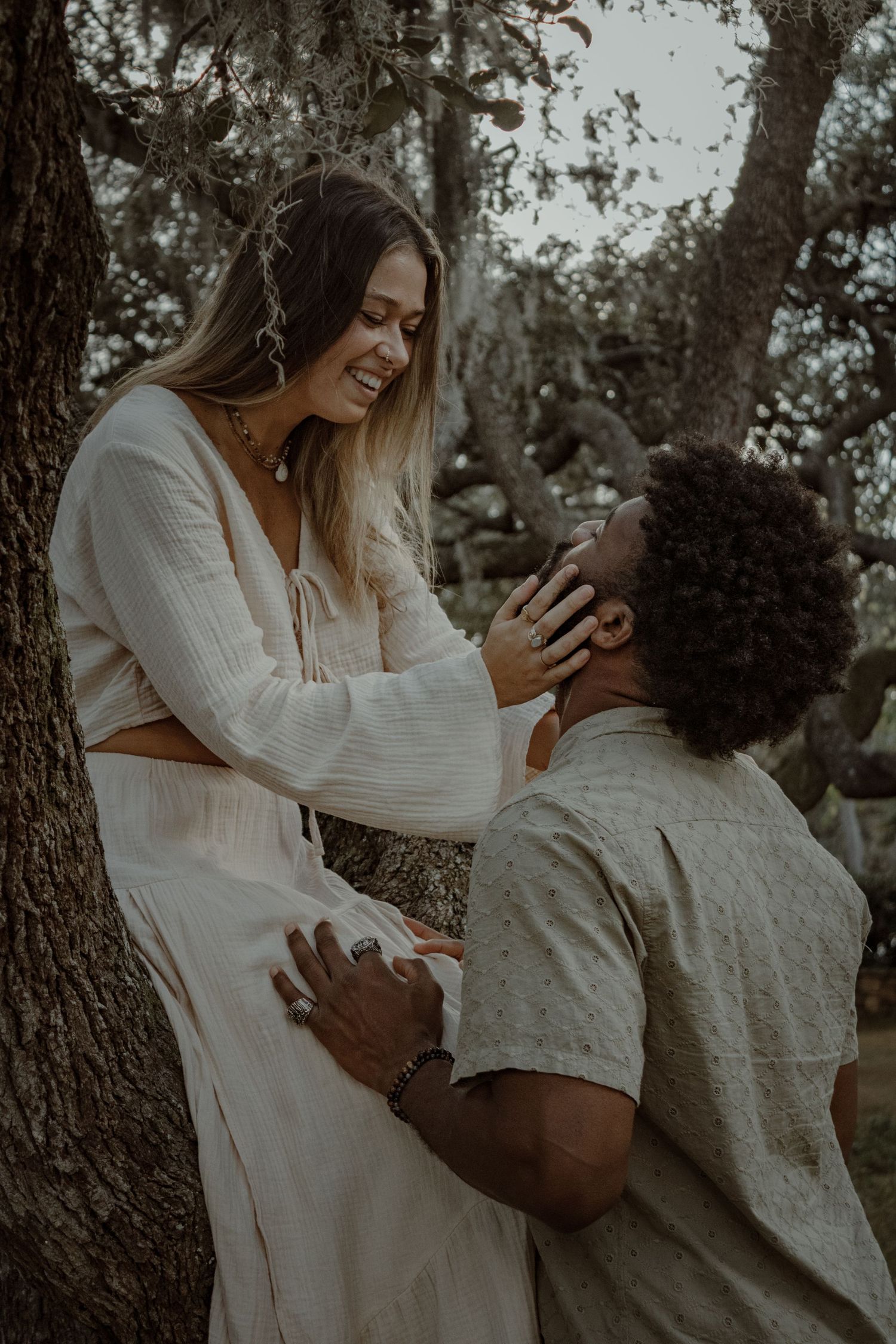 A couple shares intimate moments and laughter while sitting together outdoors against a tree.