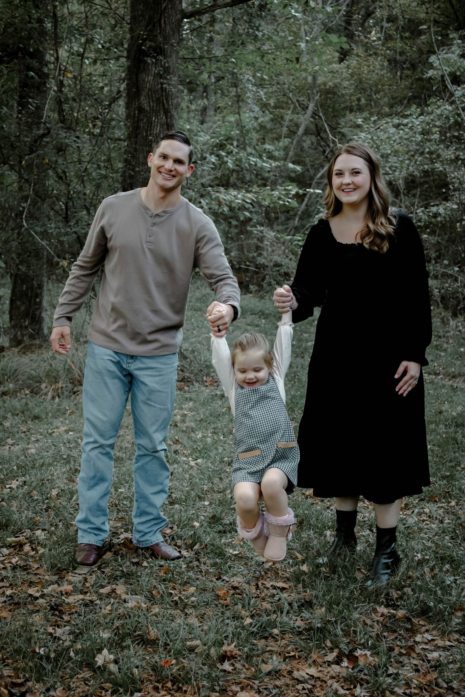 A family poses together in a wooded forest setting while swinging a small child between them.