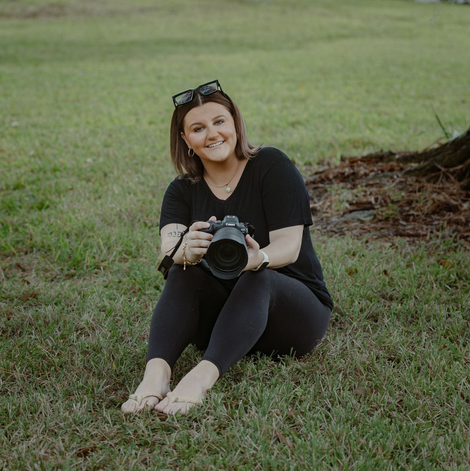 Photographer in black clothing sitting on grassy ground with professional camera in outdoor setting.