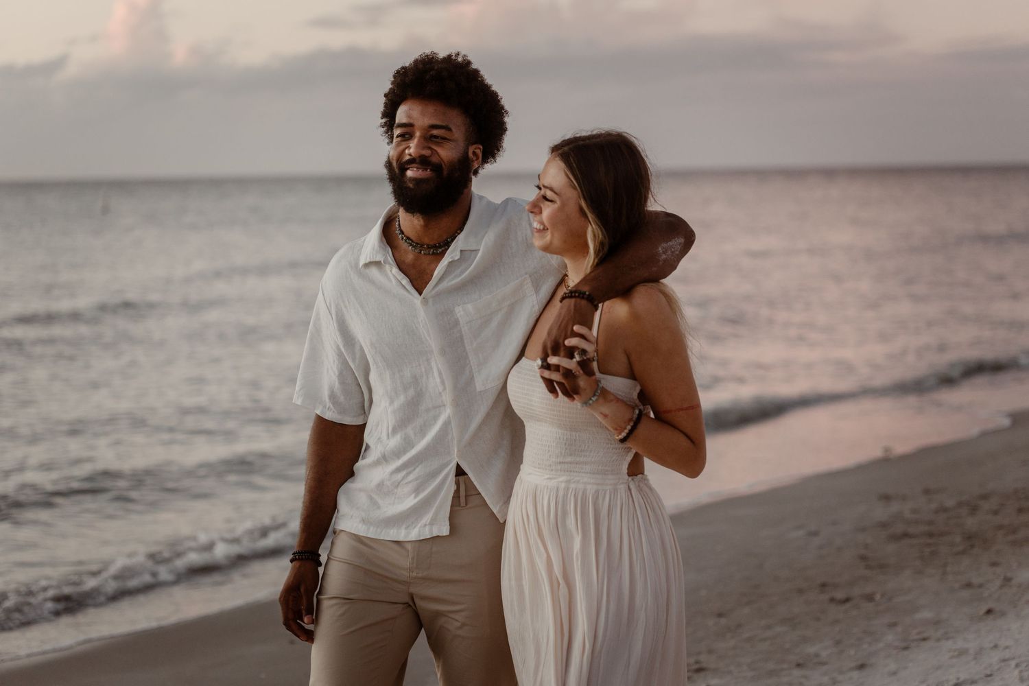 Romantic couple walking and laughing together on a sandy beach at sunset wearing white and beige outfits.