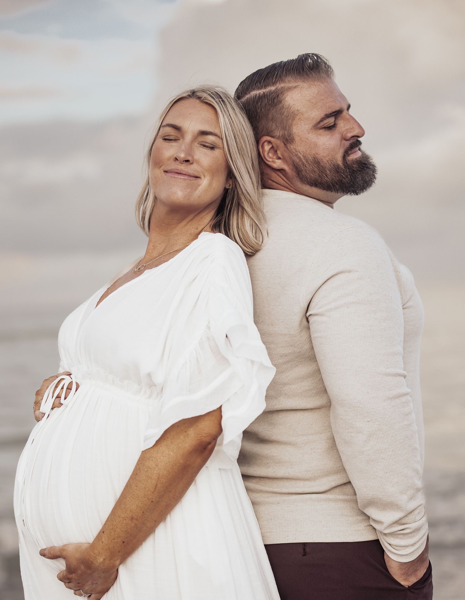 A couple in white and beige outfits pose back to back in a romantic photoshoot sequence on a cloudy day.