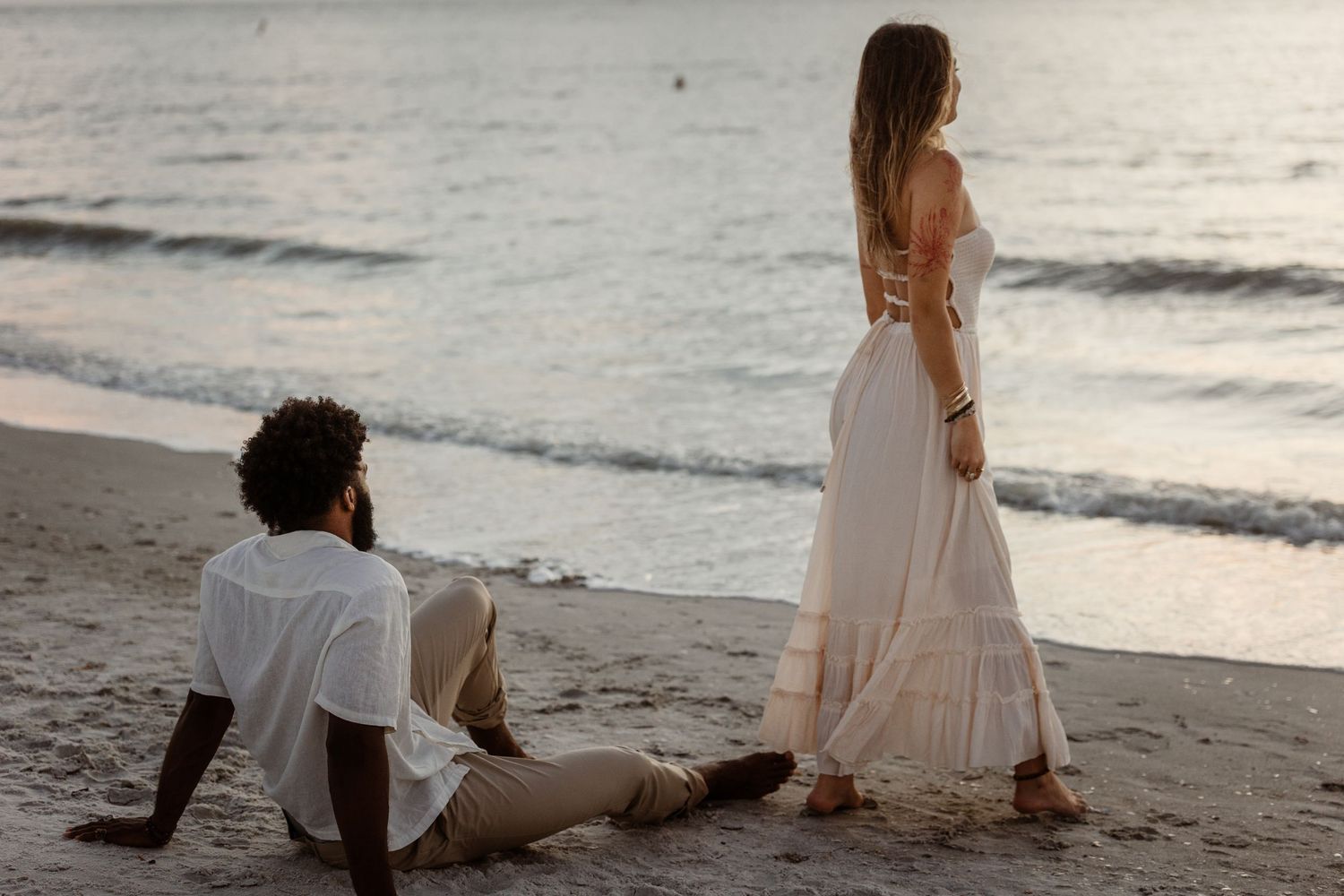A couple in light colored outfits sits together on a sandy beach at dusk with waves rolling in behind them.