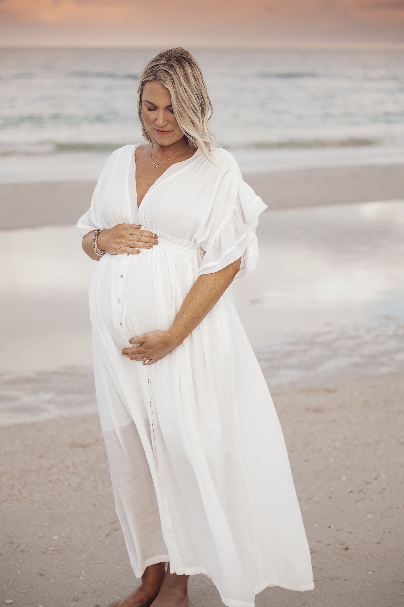A pregnant woman in a flowing white dress stands on a beach at sunset, holding her baby bump.