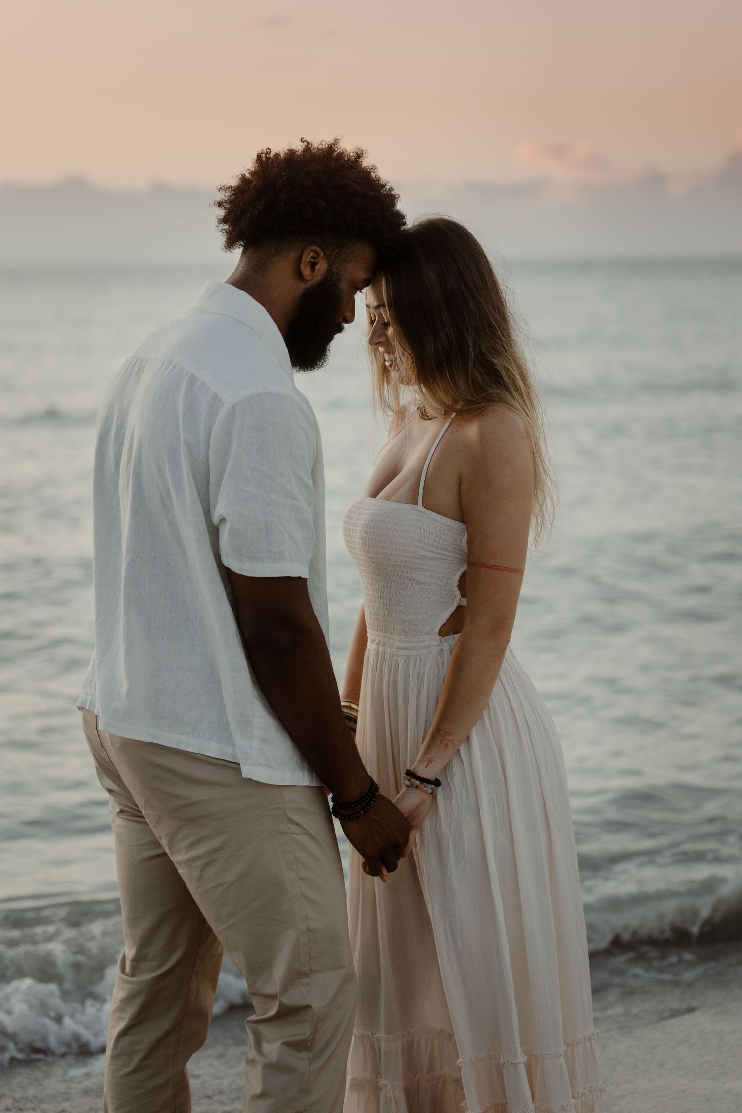 A couple in white outfits holds hands while walking along a serene beach at sunset with ocean waves in the background.