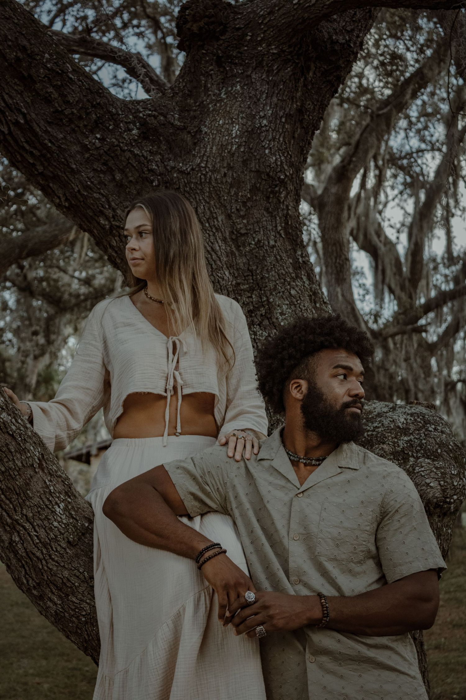 A couple poses intimately against a large oak tree in a dreamy outdoor setting.