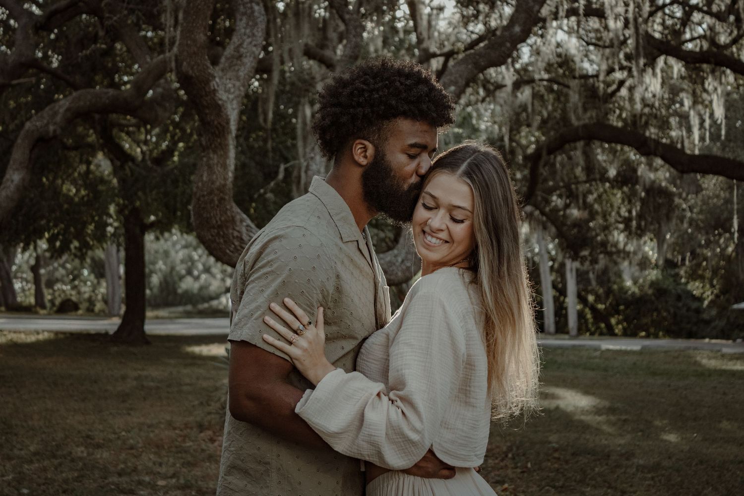 A couple shares intimate moments in a natural outdoor setting with trees and greenery in the background.