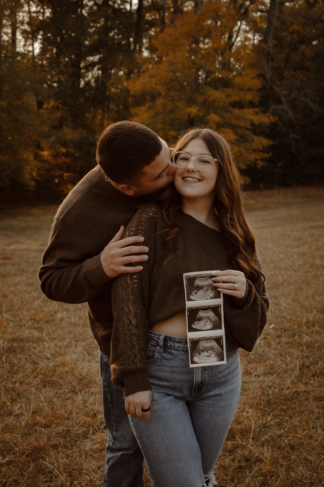 A couple embraces in a fall field while holding ultrasound photos during a pregnancy announcement photoshoot.