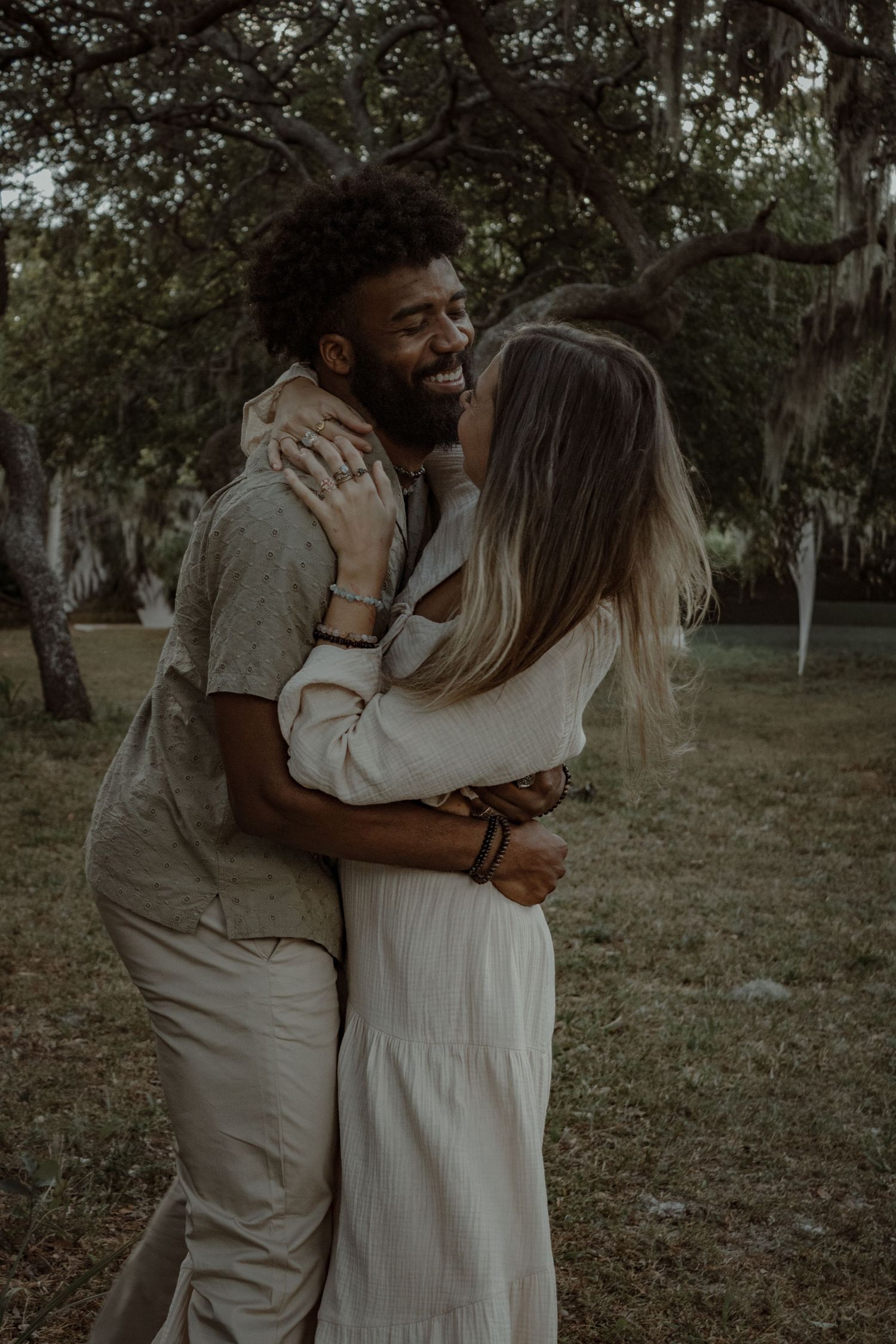 A couple shares a loving embrace in a park with Spanish moss hanging from trees.
