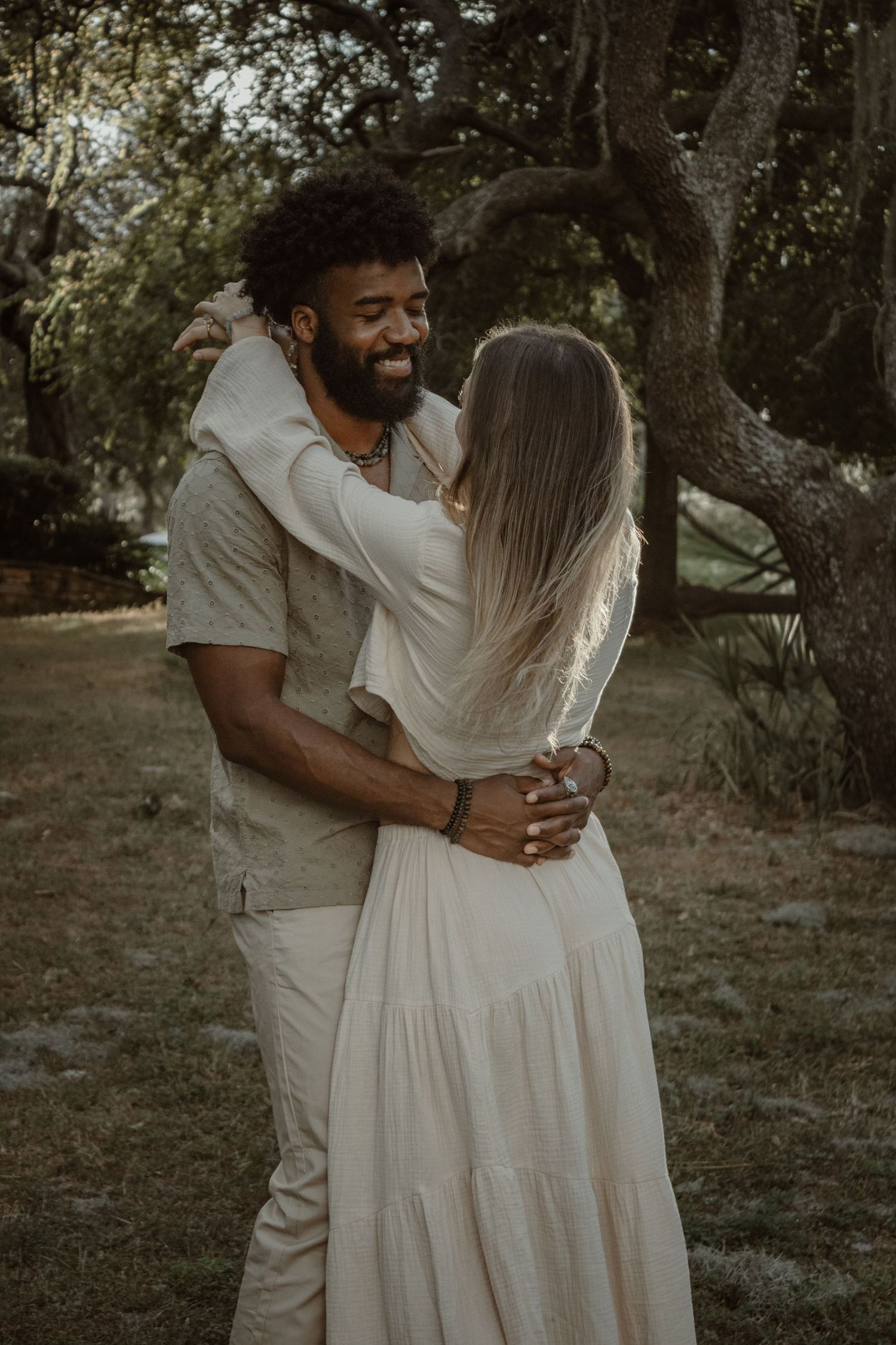A romantic couple in white outfits embrace and laugh together in a natural outdoor setting at sunset.